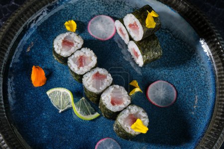 Foto de Vista superior de apetitosos rollos de maki con atún servido en plato azul con rodajas de rábano limón y flores - Imagen libre de derechos