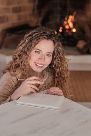 Téléchargez les photos : Femme créative joyeuse avec crayon écrit dans le journal tout en étant assis dans le salon confortable près de la cheminée à la maison - en image libre de droit