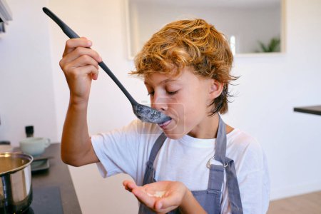 Photo for Kid in apron tasting delicious white sauce from spoon spatula during cooking process at home - Royalty Free Image