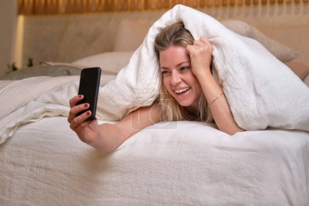 Téléchargez les photos : Femme souriante couchée sur son lit et naviguant sur son smartphone tout en se reposant dans une chambre confortable à la maison - en image libre de droit