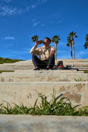 Foto de Bajo ángulo cuerpo completo de tipo hispano sentado en escalones de piedra y agua potable para refrescarse después de entrenar en monopatín en el soleado parque urbano - Imagen libre de derechos