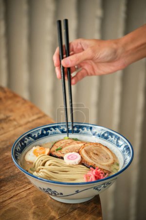 Photo for Crop anonymous person holding chopsticks while having ramen soup with noodles and fried pork in bowl - Royalty Free Image
