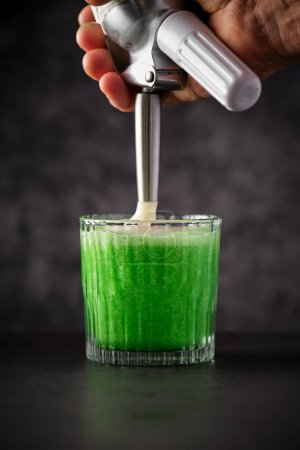 Foto de Barman masculino anónimo preparando refrescante cóctel verde y adornando con espuma batida de sifón crema en el restaurante - Imagen libre de derechos