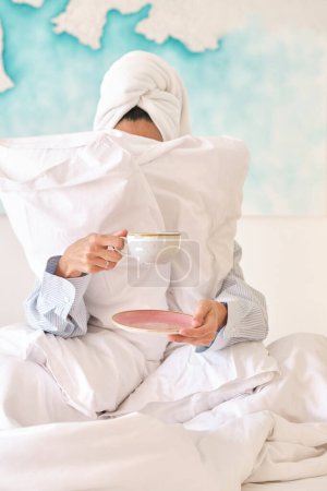 Foto de Mujer irreconocible en ropa de dormir escondida detrás de almohadas y tomando café por la mañana en la cama en el dormitorio de luz - Imagen libre de derechos