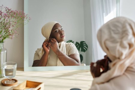 Foto de Reflejado en espejo mujer afroamericana en pañuelo beige para la cabeza poniéndose un pendiente en casa - Imagen libre de derechos