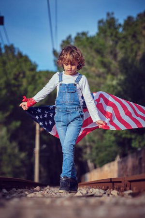 Téléchargez les photos : Garçon en jeans regardant vers le bas tout en marchant sur des pierres concassées entre les rails et tenant flotté drapeau des États-Unis - en image libre de droit