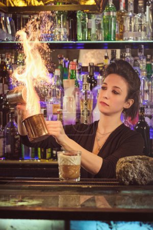 Photo for Concentrated young female bartender with dark hair sprinkling salt into metal cup and burning fire while preparing cocktail standing at counter in modern bar - Royalty Free Image