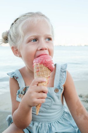 Foto de Niño de pelo justo en vestido azul mordiendo cono de helado mientras está sentado en la playa y mirando hacia otro lado pensativamente - Imagen libre de derechos