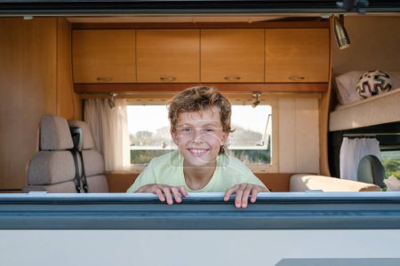 Téléchargez les photos : Garçon positif souriant et regardant la caméra tout en étant assis derrière la fenêtre du camping-car pendant les vacances d'été - en image libre de droit