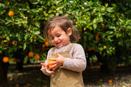 Photo for Charming child in casual apparel with ripe orange against lush green trees on summer day - Royalty Free Image