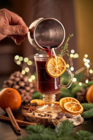 Photo for Crop anonymous person pouring alcoholic mulled wine into glass with orange slice on table with coniferous branches during Christmas holidays - Royalty Free Image