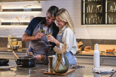 Photo for Smiling woman and man drinking wine while standing at stove and cooking together at home - Royalty Free Image