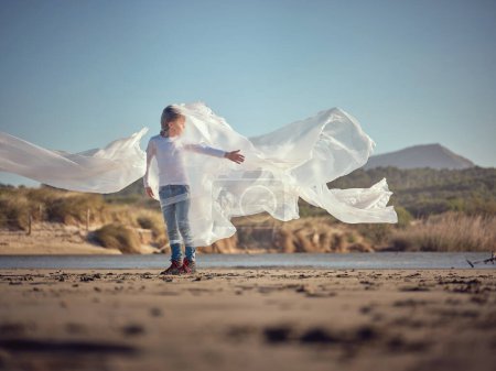 Photo for Side view of full body preteen boy rolled into transparent waving plastic waste film caught from polluted water of river standing on sandy coast - Royalty Free Image