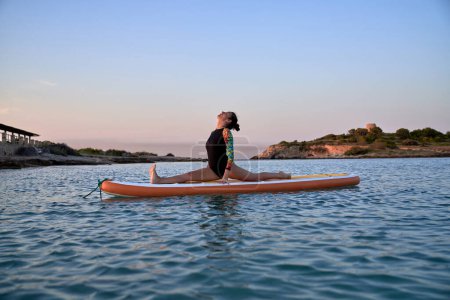 Téléchargez les photos : Vue latérale de la jeune femme en maillot de bain prenant la pose Hanumasana tout en pratiquant le yoga sur planche à pagaie au bord de la mer le soir - en image libre de droit