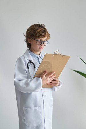Photo for Intelligent child in eyewear and white medical uniform with stethoscope and clipboard on white background - Royalty Free Image