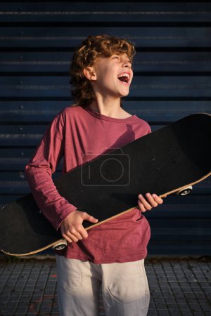 Téléchargez les photos : Joyeux petit patineur avec planche à roulettes riant et regardant loin tout en se reposant sur la rue de la ville - en image libre de droit