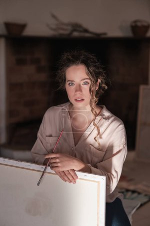 Téléchargez les photos : Femme talentueuse avec pinceau professionnel assis près de la toile et regardant la caméra tout en peignant dans un studio moderne - en image libre de droit