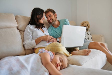 Photo for Happy man caressing tummy of pregnant woman with netbook and sitting on sofa near napping baby girl in living room at home - Royalty Free Image