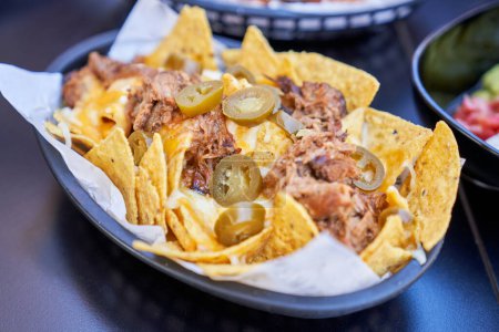 Téléchargez les photos : Du dessus de la portion de nachos de maïs savoureux garnie de tranches de jalapeno de viande tirée et de sauce dans un panier noir - en image libre de droit