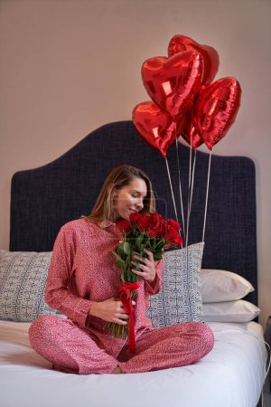 Téléchargez les photos : Heureuse femme assise avec des jambes croisées sur le lit avec un bouquet de ballons en aluminium en forme de coeur et profitant d'un arôme de bouquet de roses rouges fraîches en fleurs - en image libre de droit