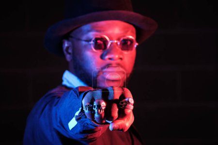Foto de Hombre étnico en gafas y sombrero mirando a la cámara mientras extiende el puño con anillos en enfoque selectivo sobre fondo oscuro - Imagen libre de derechos