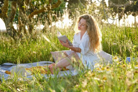 Photo for Full body sensitive young female wearing white sundress reading interesting book while resting on blooming abundant lawn in sunny summer countryside - Royalty Free Image