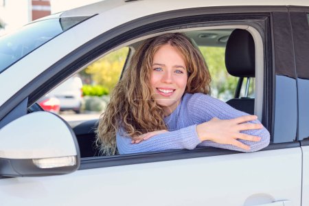 Photo for Cheerful young female driver in casual wear sitting in car parked in sunny city street and looking at camera with pretty smile - Royalty Free Image