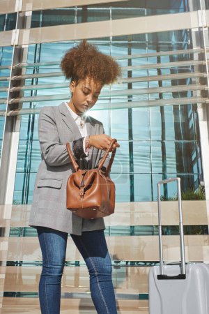 Photo for Low angle of young African American female traveler in smart casual outfit with luggage standing near glass wall of airport building in city - Royalty Free Image