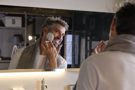 Téléchargez les photos : Homme souriant debout dans la salle de bain moderne près du miroir et regardant la réflexion tout en appliquant de la mousse à raser et en faisant la routine de soins de la peau du matin à la maison - en image libre de droit