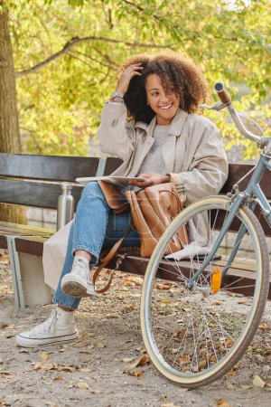 Téléchargez les photos : Corps complet de jeune femme ethnique élégante positive reposant avec un livre sur un banc en bois près du vélo dans le parc et touchant les cheveux - en image libre de droit