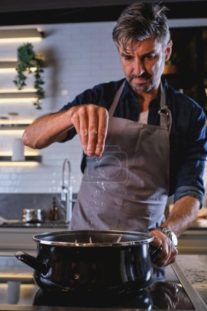Foto de Hombre barbudo en delantal gris poniendo sal en la cacerola mientras se prepara la comida en la cocina moderna - Imagen libre de derechos
