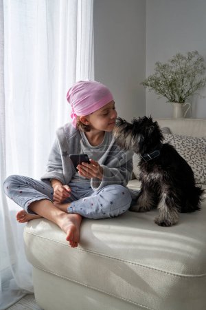 Téléchargez les photos : Pleine longueur de l'enfant pieds nus dans le bandeau rose en utilisant le téléphone portable tout en passant du temps avec un petit chien dans le salon - en image libre de droit