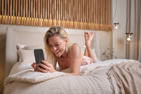 Foto de Cuerpo completo de mujer positiva en camisón de seda divirtiéndose mientras lee el mensaje de texto en el teléfono inteligente y descansando en el acogedor dormitorio en casa - Imagen libre de derechos