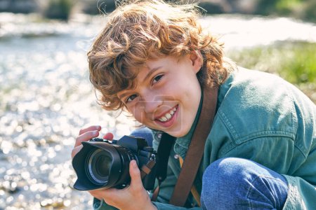 Photo for Happy boy taking picture with professional photo camera and looking at camera while sitting on haunches near flowing river on grassy shore - Royalty Free Image