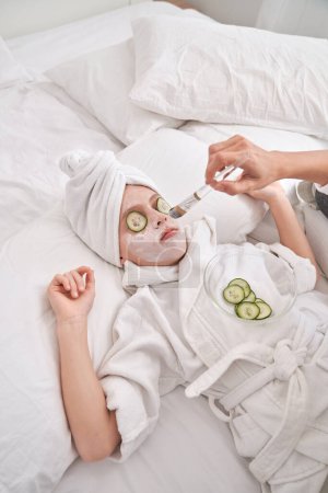 Photo for From above of crop anonymous mother applying moisturizing mask with brush on face of kid in bathrobe relaxing on comfortable bed with cucumber slices on eyes during spa session - Royalty Free Image