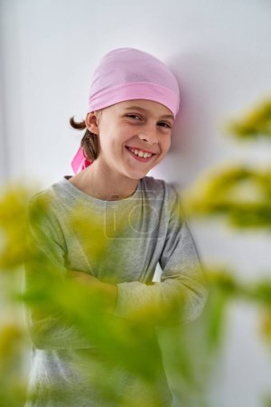 Photo for Positive preteen unhealthy boy in casual clothes and bandana on head smiling leaning on white wall and looking at camera - Royalty Free Image