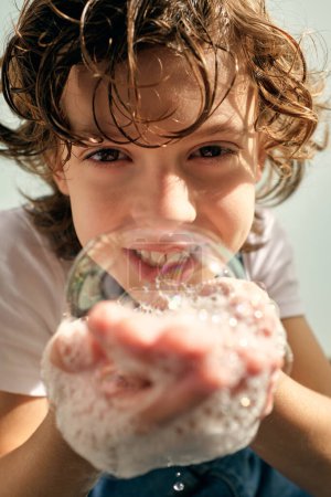 Photo for Portrait of smiling adorable kid with wet curly hair in casual clothes holding soap bubble on palms and looking at camera - Royalty Free Image