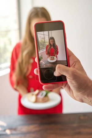 Photo for Crop anonymous person taking photo of young woman in red dress with delicious tartar while standing in kitchen - Royalty Free Image