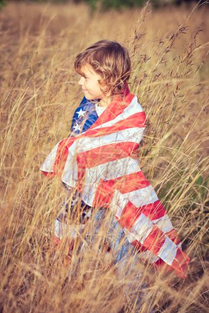 Foto de Vista lateral del lindo niño preadolescente envuelto en la bandera nacional de Estados Unidos de América de pie en el prado de centeno - Imagen libre de derechos