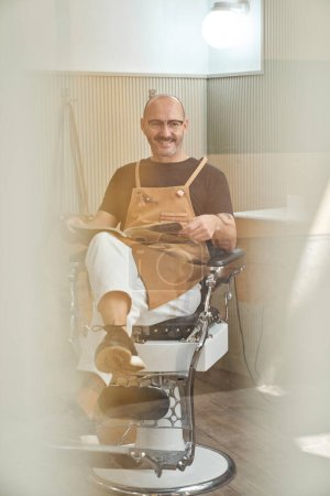 Foto de Peluquero masculino sonriente con bigote en delantal sentado en silla y leyendo revista mientras descansa en el salón de peluquería - Imagen libre de derechos