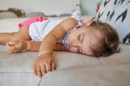 Téléchargez les photos : Bébé fille insouciante couchée sur un canapé confortable et faisant la sieste dans le salon à la maison le jour - en image libre de droit