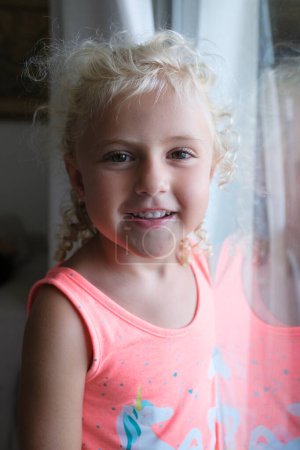 Photo for Portrait of adorable little girl with blond hair looking at camera while standing near window at home - Royalty Free Image