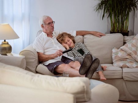 Téléchargez les photos : Corps complet de grand-père concentré avec petit-fils regardant un film intéressant tout en étant assis sur un canapé confortable dans le salon à la maison - en image libre de droit
