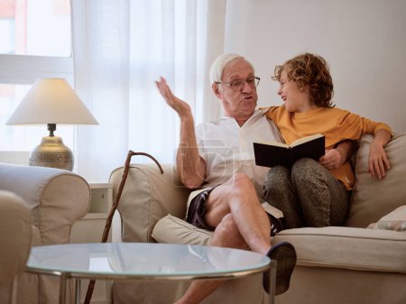 Foto de Muchacho sonriente en ropa casual mirando al abuelo mientras leen el libro juntos en un cómodo sofá en la sala de estar ligera en casa - Imagen libre de derechos