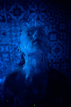 Photo for Kid with closed eyes under water fluid in bathroom with ornamental tiled wall in blue neon light - Royalty Free Image