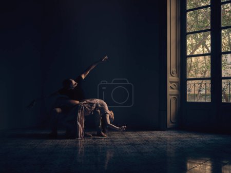Foto de Cuerpo completo de bailarina acostada sobre las rodillas del bailarín masculino mientras realiza el movimiento de danza de ballet en habitación oscura con ventana - Imagen libre de derechos