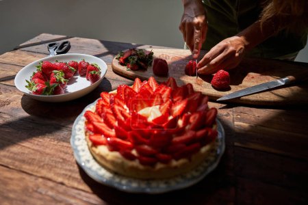 Foto de Alto ángulo de persona anónima cortando frutas frescas maduras de fresa con cuchillo mientras que la torta decorada colocada a la luz del sol en la mesa de madera - Imagen libre de derechos