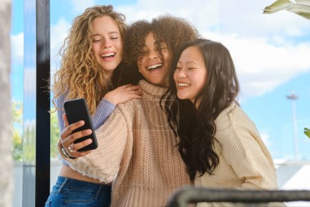 Téléchargez les photos : Joyeuses copines multiraciales souriant et prenant autoportrait sur téléphone portable tout en se tenant contre le ciel bleu dans la journée ensoleillée - en image libre de droit