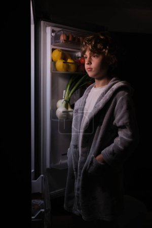 Photo for Hungry boy in nightwear standing near opened refrigerator with various fresh products while choosing food in dark kitchen at home - Royalty Free Image