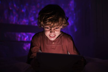 Photo for Irritated preteen child in sleepwear playing game on tablet while sitting on bed in dark bedroom at night time in apartment - Royalty Free Image